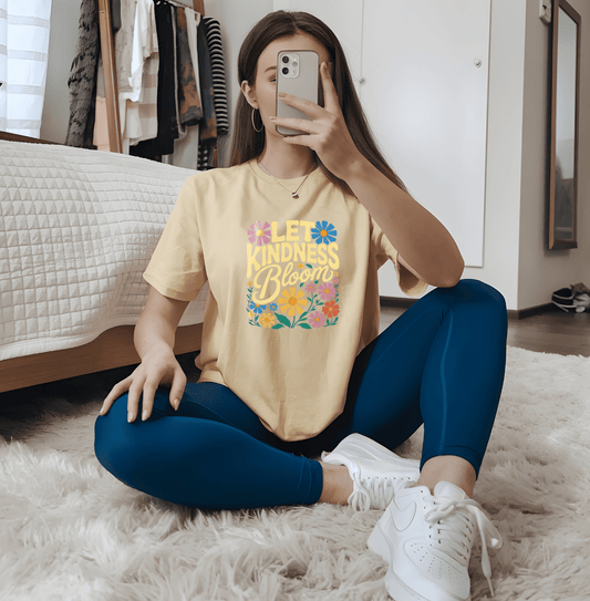 A person sits cross-legged on the floor in a bedroom, taking a mirror selfie. They wear the Let Kindness Bloom yellow T-shirt by DB Custom Creations, featuring colorful flowers and vibrant DTF Transfer prints, paired with blue leggings.