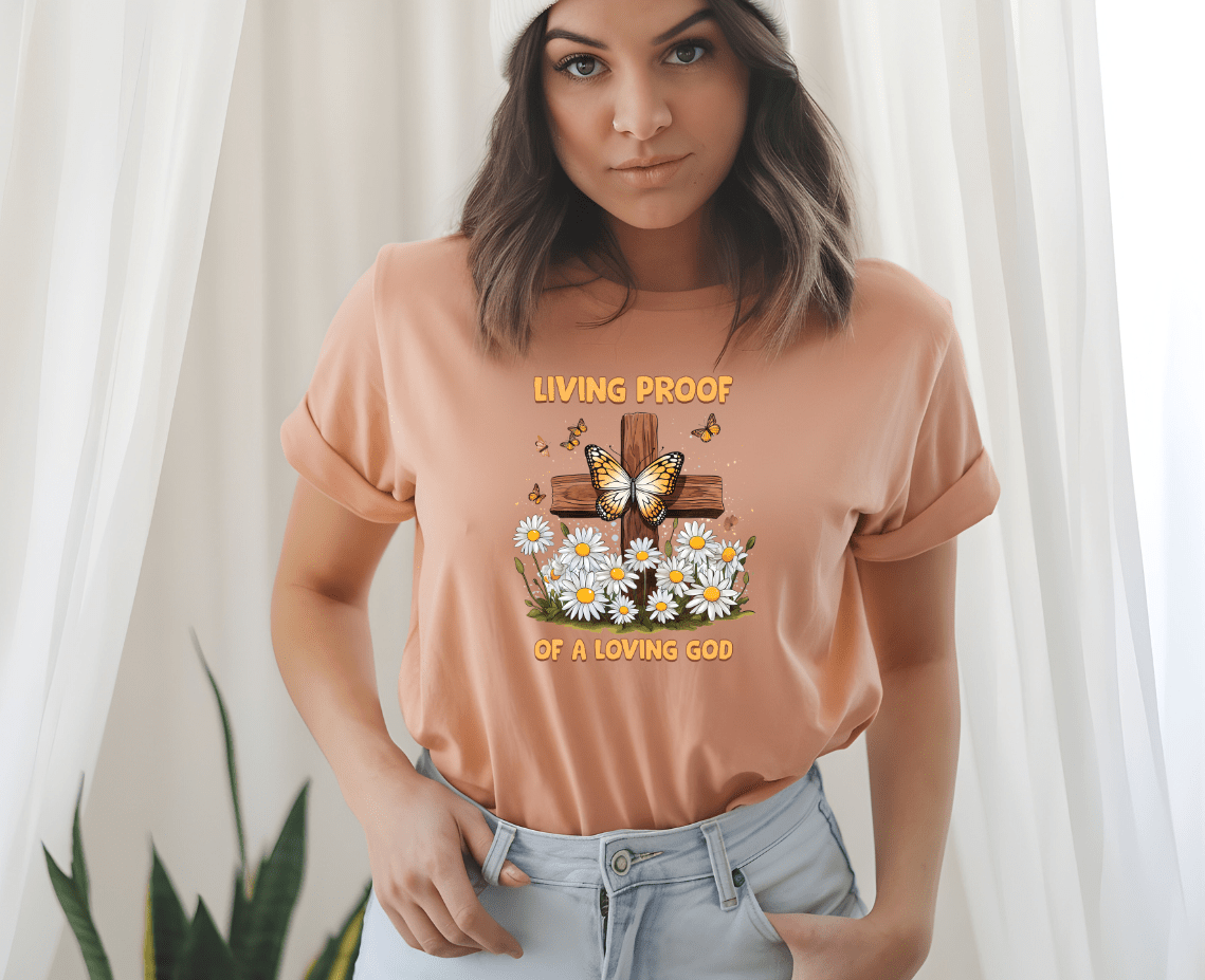 A woman in a peach Living proof of a loving God t-shirt by DB Custom Creations, with DTF printing and a cross, butterfly, and daisies design, stands indoors near white curtains and a green plant.