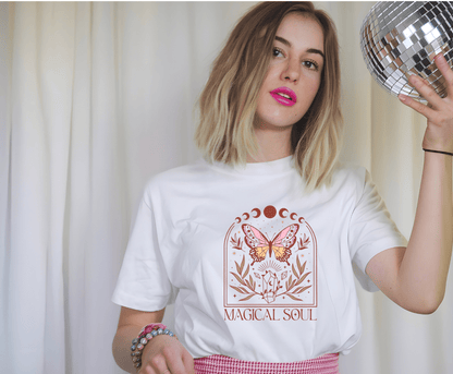 A young woman with a blonde bob and pink lipstick holds a disco ball, wearing the Magic Soul t-shirt by DB Custom Creations, featuring a mystical butterfly and flower design in 11-inch DTF print, plus pastel bead bracelets against white curtains.