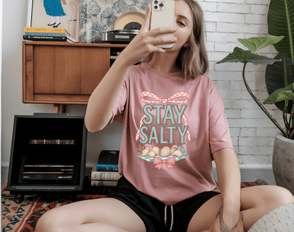 A woman sits on the floor taking a selfie, wearing DB Custom Creations stay salty pink t-shirt featuring a seashell and ribbon DTF Transfer design, in a cozy room with books, plants, and a record player.