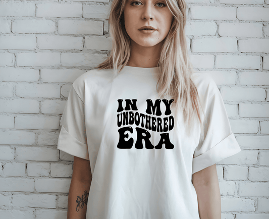A woman with long blonde hair stands by a white brick wall, wearing DB Custom Creations’ unbothered era white T-shirt, featuring bold black “IN MY UNBOTHERED ERA” text printed via DTF.