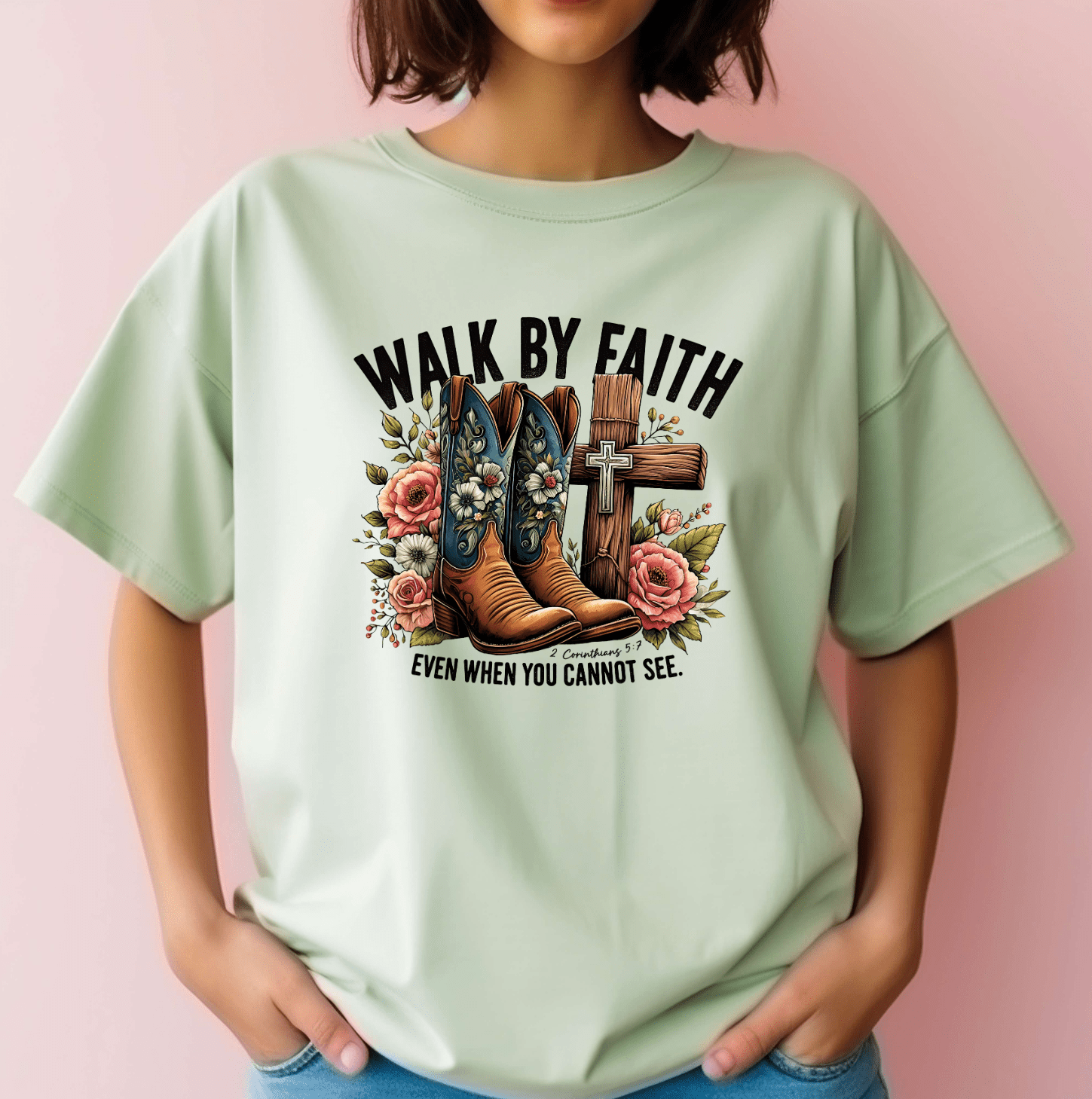 A person with short brown hair stands against a pink background, wearing the Walk by Faith light green tee from DB Custom Creations, featuring DTF-printed cowboy boots, flowers, a cross, and the phrase WALK BY FAITH EVEN WHEN YOU CANNOT SEE.
