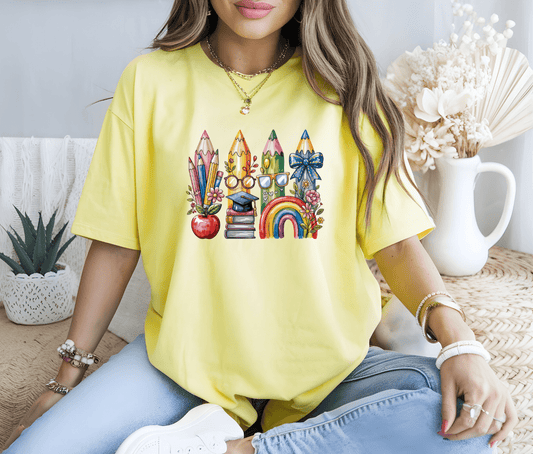 A woman in a yellow watercolor back to school t-shirt by DB Custom Creations—decorated with DTF prints of pencils, books, an apple, glasses, a rainbow, and a bow—sits cross-legged on a woven seat near dried flowers and a plant.