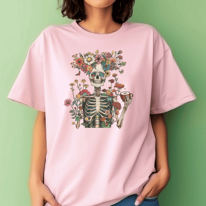 A person wears DB Custom Creations’ Whimsical Skeleton light pink tee, featuring a skeleton with a pipe and colorful flowers in 11-inch vibrant DTF print. The background is a plain light green wall.