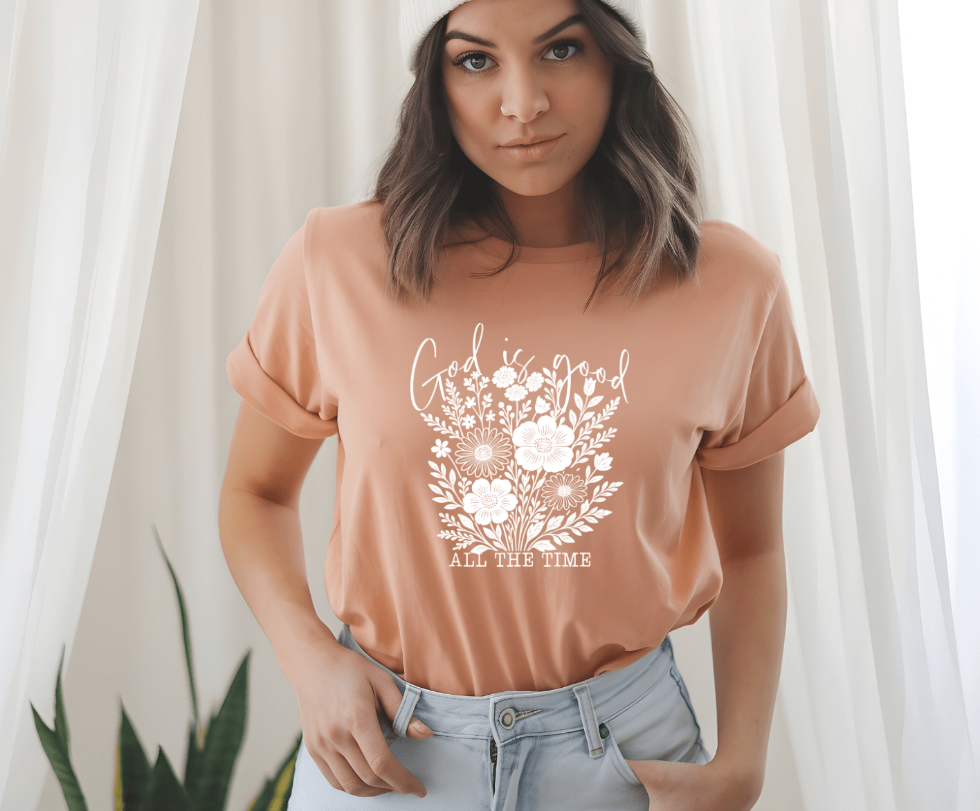 A woman in light blue jeans and a knit beanie wears the DB Custom Creations God is good white floral peach t-shirt, featuring a white floral design and the words “God is good all the time,” standing indoors and looking at the camera.