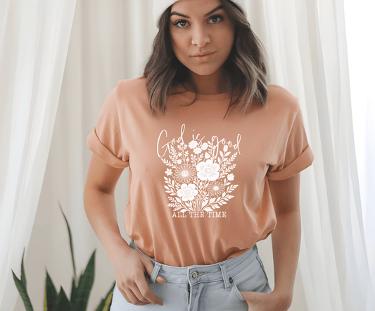 A woman in light blue jeans and a knit beanie wears the DB Custom Creations God is good white floral peach t-shirt, featuring a white floral design and the words “God is good all the time,” standing indoors and looking at the camera.